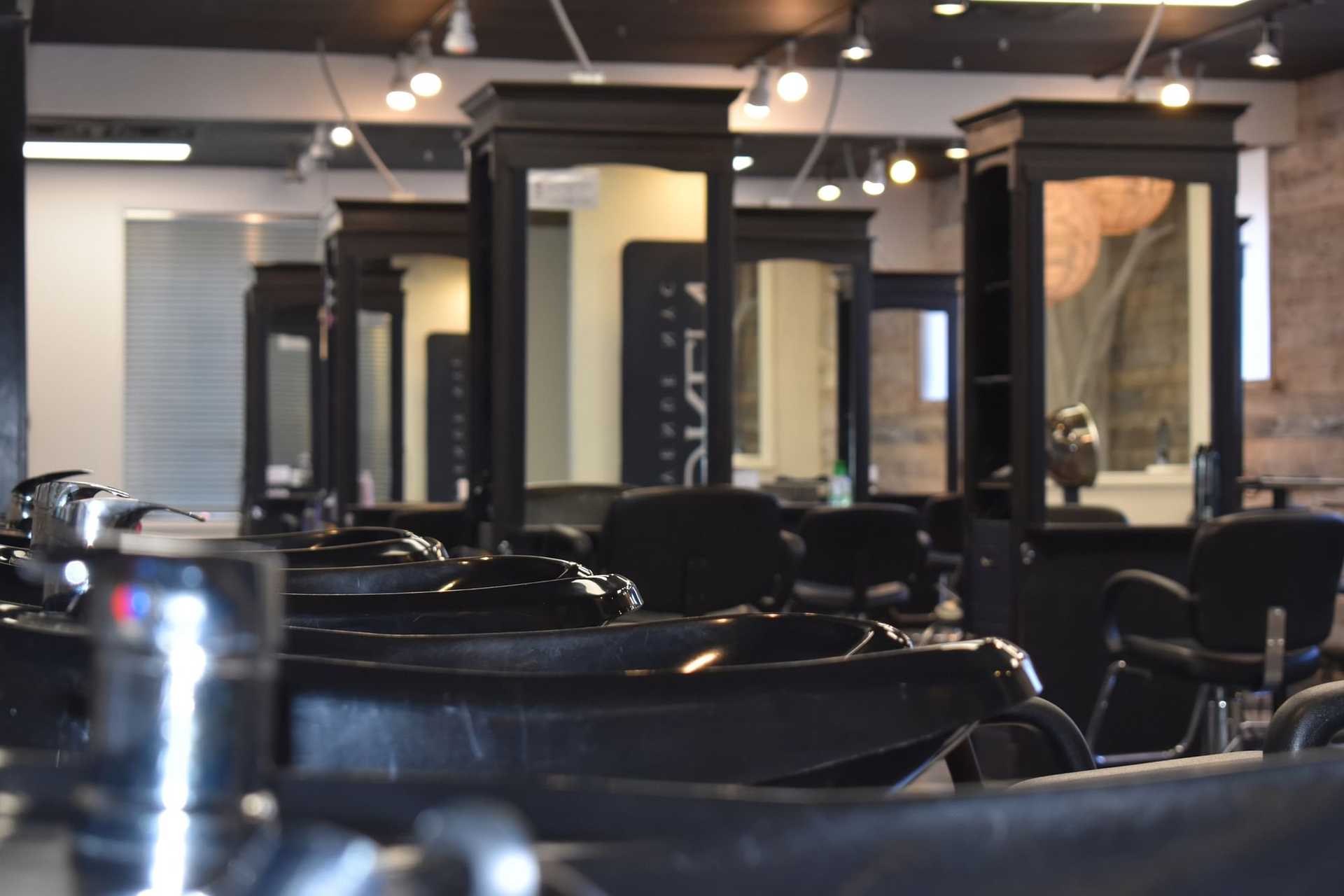 Empty salon with black chairs and washing stations, mirrors, and light fixtures in the background.